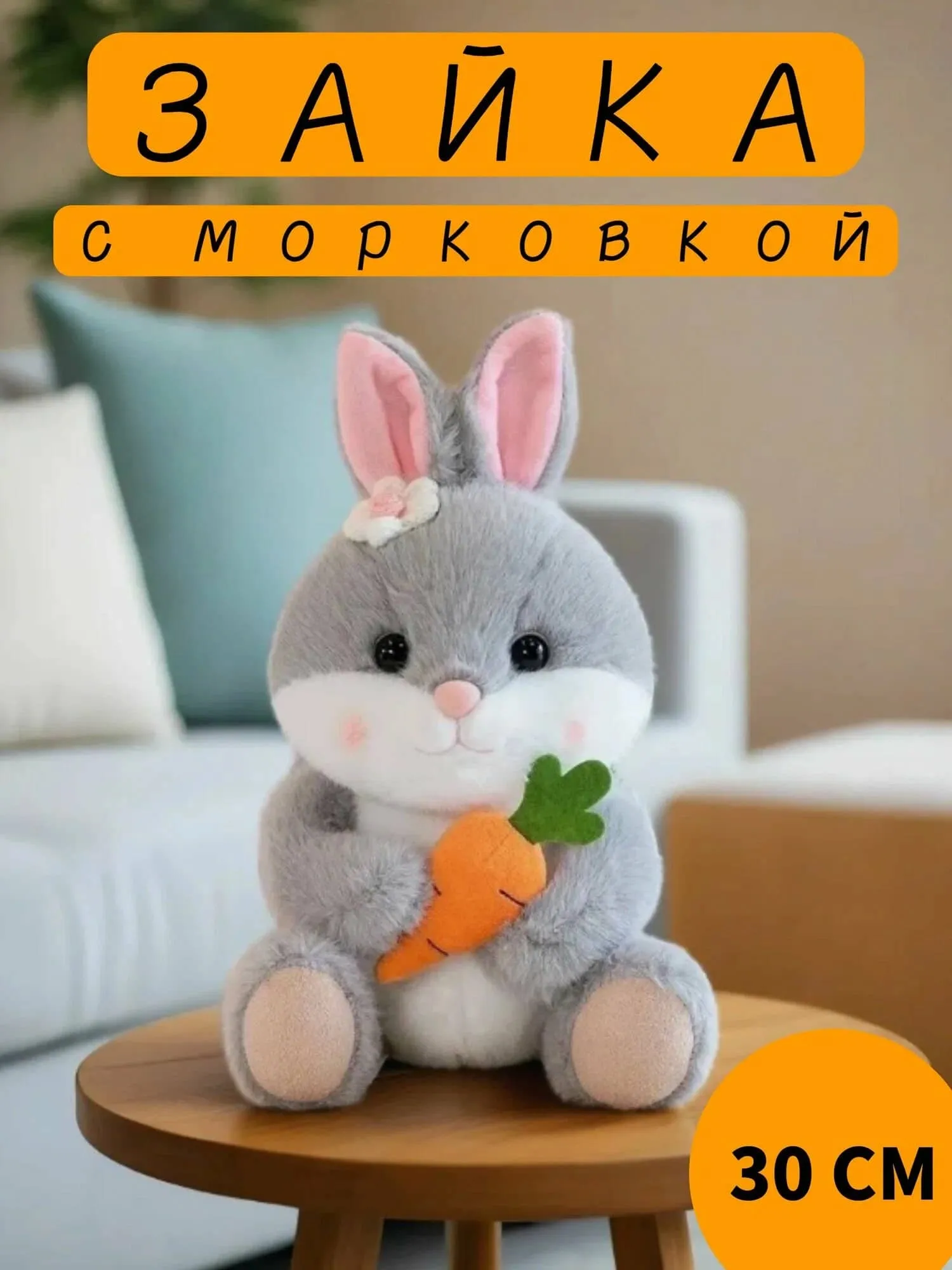 Милый серый плюшевый зайчик с морковкой в лапках, розовые ушки, цветочек на голове, размер 30 см, сидит на деревянной поверхности